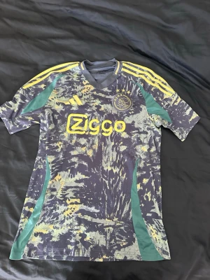 Ajax x Adidas matchtröja Ziggo M - Ajax matchtröja från Adidas i storlek M. Tröjan har ett unikt mönster i blått, gult och grönt med gula detaljer och Ziggo-logga på bröstet. Klassiska Adidas-ränder på axlarna och Ajax-emblem på vänster bröst. Tillverkad i lätt funktionsmaterial.
