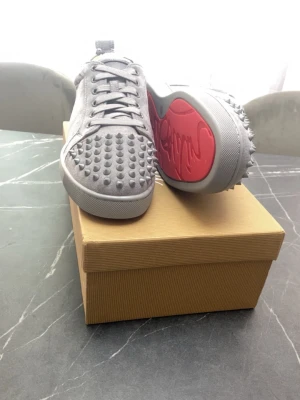Gråa Christian Louboutin sneakers - Säljer ett par gråa sneakers från Christian Louboutin med ikoniska röda sulor och coola nitar över hela ovandelen. Skorna har en låg profil och är tillverkade i slätt läder som ger en lyxig känsla. Perfekta för dig som vill sticka ut med din stil.