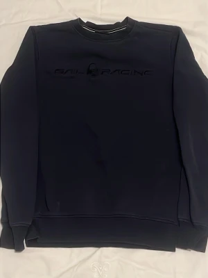 Mörkblå sweatshirt Sail Racing - Mörkblå sweatshirt från Sail Racing med diskret logga broderad på bröstet. Klassisk rund halsringning och långa ärmar. Perfekt för en avslappnad och stilren look.