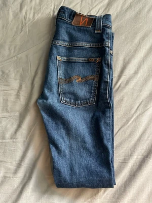 Blå jeans från Nudie Jeans  - Säljer ett par klassiska blå jeans från Nudie Jeans med snygga kontrastsömmar och ikonisk våg på bakfickan. Jeansen  passformen slim fit. Pris kan diskuteras vid snabb affär.