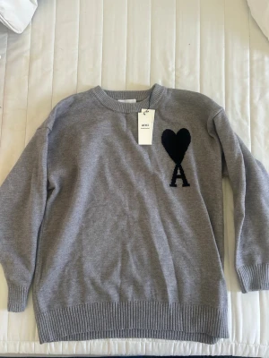 Grå stickad tröja från Ami Paris - Snygg grå stickad tröja från Ami Paris med svart hjärta och bokstaven A broderat på bröstet. Tröjan har rund halsringning, ribbade muddar och är tillverkad i mjukt material som passar perfekt till höst och vinter.