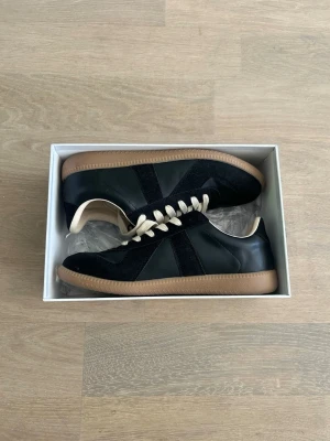 Maison Margiela svarta sneakers - Snygga svarta sneakers från Maison Margiela med beige snörning och gummisula. Skorna har en mix av läder och mocka, klassisk siluett och diskret design. Perfekta för dig som gillar stilrena och exklusiva sneakers. Pris kan diskuteras!