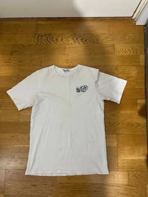Vit Moncler t-shirt med broderad logga - Vit t-shirt från Moncler i bomull med rund hals och korta ärmar. På bröstet sitter två broderade Moncler-loggor i vitt, rött och blått. Enkel och stilren design som passar perfekt till jeans eller shorts.