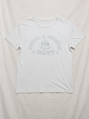 Ralph Lauren Denim & Supply - Vit t-shirt från Denim & Supply Ralph Lauren med tryck framtill som säger 'NAVY' och har ett marint motiv. Klassisk rund halsringning och korta ärmar. Tillverkad i mjuk bomull för en skön känsla.