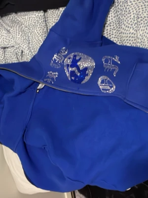 Aimforthemoon Herr - Säljer en blå hoodie med huva och dragkedja. På framsidan finns silvriga tryck med olika symboler och text, samt en stor ficka framtill. Perfekt för dig som gillar streetwear och vill sticka ut med coola detaljer.