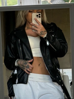 Svart skinnjacka med bälte - Snygg svart skinnjacka med klassisk bikerstil, dragkedjor och bälte i midjan. Jackan har breda slag och silvriga detaljer som ger en cool look. Perfekt för dig som vill ha en edgy stil och gillar streetmode. Aldrig använd plasten är kvar på dragkedjor