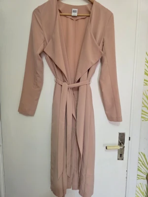 Ljusrosa trenchcoat från Vero Moda - Stilren ljusrosa trenchcoat från Vero Moda med långa ärmar, bred krage och knytskärp i midjan. Jackan har en öppen front och är tillverkad i ett mjukt, tunt tyg som ger ett snyggt fall. Perfekt för dig som vill ha en trendig och enkel look.