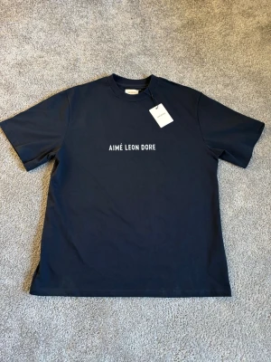 Mörkblå Aimé Leon Dore t-shirt - Snygg mörkblå t-shirt från Aimé Leon Dore med vit logotyp på bröstet. Klassisk passform med rund hals och korta ärmar. Tillverkad i mjuk bomull som känns skön mot huden. Perfekt för dig som gillar stilrena och trendiga plagg.