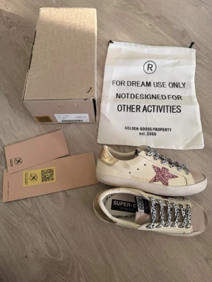 Golden Goose sneakers med glitterstjärna - Snygga sneakers från Golden Goose i beige läder med guldiga detaljer och en rosa glitterstjärna på sidan. Skorna har svarta och vita snören med mönster och klassisk låg siluett. Perfekta för dig som vill sticka ut med en unik och trendig look.