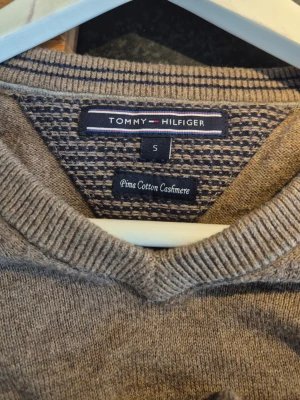 Beige stickad tröja Tommy Hilfiger - Beige stickad tröja från Tommy Hilfiger i mjuk prima cotton cashmere-blandning. V-ringad modell med diskret logga broderad på bröstet och ribbade muddar vid ärmslut och nederkant. Perfekt för en stilren och avslappnad look.