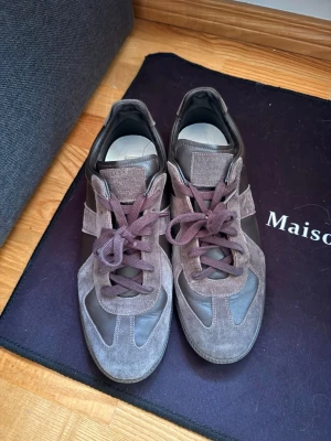    Maison Margiela - Snygga sneakers från Maison Margiela i gråbrunt mocka och läder. Klassisk design med paneler och matchande snörning. Perfekta för dig som gillar stilrena och trendiga skor med en lyxig känsla.