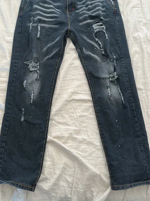 Mörkblå slitna jeans med raka ben - Säljer ett par mörkblå jeans med tydliga slitningar och hål på framsidan. Jeansen har även små färgstänk.