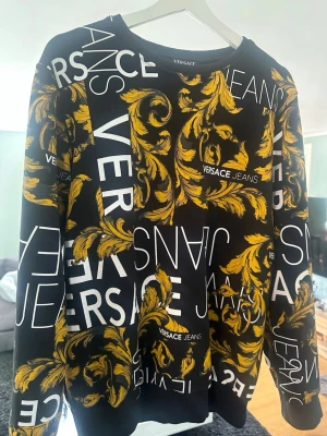 Svart tröja med mönster - Svart tröja med stort vitt logotryck och barockinspirerat gult mönster över hela plagget. Långärmad modell med rund halsringning. Perfekt statement-piece för dig som gillar lyxig streetstyle.