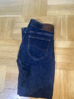 Mörkblå Lee jeans med rak passform - Snygga mörkblå jeans från Lee med klassisk rak passform och femficksdesign. Jeansen har tydliga kontrastsömmar och en brun läderpatch med Lee-loggan bak i midjan. Perfekta för en avslappnad och stilren look.