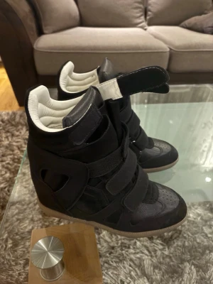 Isabel Marant storlek 38 - Säljer ett par svarta high top sneakers med cool design och flera kardborreband. Skorna har en chunky sula i beige och detaljer i både mocka och läderimitation. Insidan är vit och vadderad för extra komfort. Perfekta för dig som vill sticka ut med din stil.