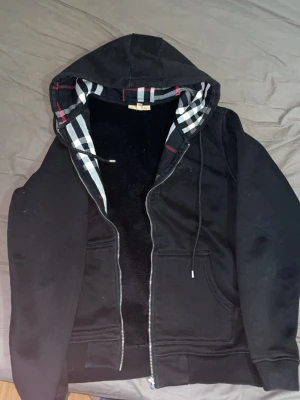 Svart hoodie med rutigt foder - Snygg svart hoodie med dragkedja och huva. Insidan av huvan har ett klassiskt rutigt mönster i svart, vitt och rött. Hoodien har fickor framtill och justerbara snören i huvan. Perfekt för en avslappnad och trendig stil.