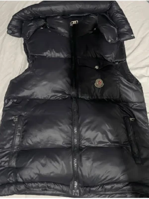 Svart dunväst från Moncler - Snygg svart dunväst från Moncler med glansig finish och hög krage. Västen har dragkedja framtill, två sidofickor med dragkedja och en bröstficka med knapp. Klassisk Moncler-logga på bröstet och diskret rödvit rand på insidan av kragen. Perfekt för lager-på-lager.