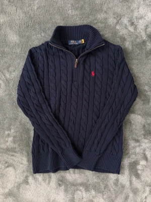 Mörkblå kabelstickad tröja Polo Ralph Lauren - Mörkblå kabelstickad tröja från Polo Ralph Lauren med halv dragkedja och röd broderad logga på bröstet. Tröjan har hög krage och ribbade muddar vid ärmslut och nederkant. Perfekt för lager på lager och klassisk stil.