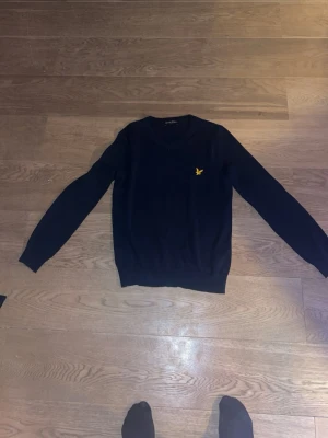 Mörkblå stickad tröja Lyle & Scott - Mörkblå stickad tröja från Lyle & Scott med klassisk gul logga på bröstet. Tröjan har V-ringning och långa ärmar, perfekt för lager på lager. Enkel och stilren design som passar till det mesta.på andra bilden är spegeln smutsig, har aldrig använt och den är small