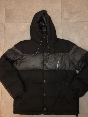 Svart pufferjacka från Adidas - Säljer en svart pufferjacka från Adidas med huva och dragsko. Jackan har en blank panel över bröstet med Adidas-logga och tre diskreta ränder. Fylld för extra värme och har knappar framtill samt elastiska muddar vid ärmsluten. Perfekt för kalla vinterdagar.