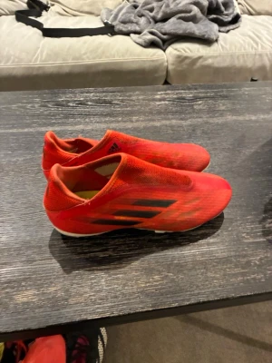 Röda Adidas fotbollsskor utan snörning - Säljer ett par röda Adidas fotbollsskor med svarta detaljer och vita dobbar. Skorna är utan snörning och har en strömlinjeformad design för bästa bollkontroll. Ovandelen är i syntetmaterial och sulan är anpassad för spel på gräs och konstgräs:)
