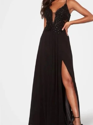 Svart långklänning med slits - Elegant svart långklänning med glittrig överdel, smala axelband och djup v-ringning. Kjolen är flödande med hög slits framtill som ger en snygg touch. Perfekt för dig som vill sticka ut på bal eller fest.uppsyd 10cm så passar någon som är 165cm ungefär om man bär klackar 