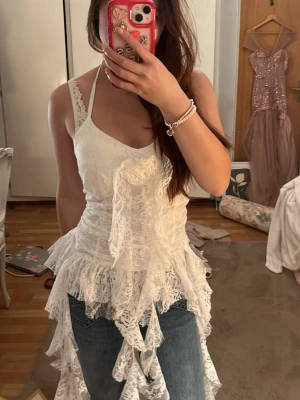 Vit spetslinnetopp med volanger - Romantisk vit linnetopp i spets med volangdetaljer och dekorativ rosett framtill. Toppen har smala axelband och asymmetrisk nederkant som ger en bohemisk känsla. Perfekt för dig som vill sticka ut med en unik och feminin stil.