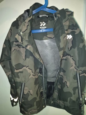  Barn Kamo vindjacka från 157 Funk stl 134-140 - Snygg vindjacka i grönt kamouflagemönster från 157 Funk. Jackan har huva, dragkedjor framtill och reflexdetaljer på ärmen. Perfekt för dig som vill ha en streetig och funktionell look. Meshfoder på insidan för extra komfort.