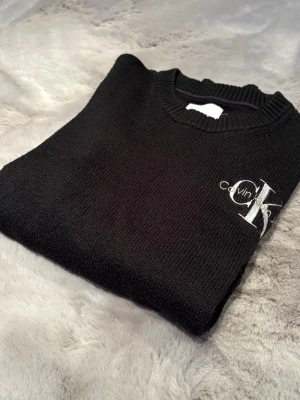 Svart stickad tröja Calvin Klein - Svart stickad tröja från Calvin Klein Jeans med rund halsringning och ribbade muddar. Tröjan har en stor vit CK-logga broderad på bröstet och är gjord i ett mjukt, tjockt material. Perfekt för dig som gillar stilrena och klassiska plagg. Den passar, S - 170.
