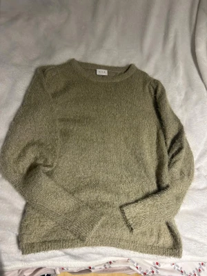 Beige stickad tröja från Vila - Mysig beige stickad tröja från Vila med rund halsringning och långa ärmar. Tröjan har en fluffig, mjuk struktur och ribbstickade muddar vid ärmslut och nederkant. Perfekt för lager på lager och enkel att matcha med jeans eller kjol.