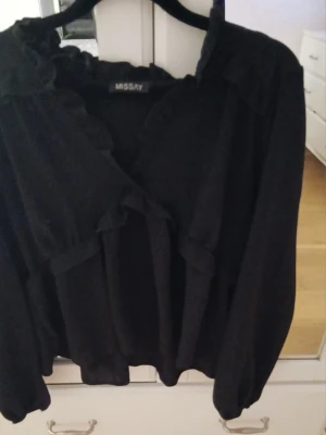 Svart volangblus från Missay - Snygg svart blus från Missay med volangdetaljer längs krage och framstycke. Blusen har v-ringning och långa ärmar med lätt puff. Tillverkad i ett tunt, mjukt tyg som ger ett flowy intryck. Perfekt för dig som gillar en chic och trendig look. Kan skicka fler bilder vid förfrågan 