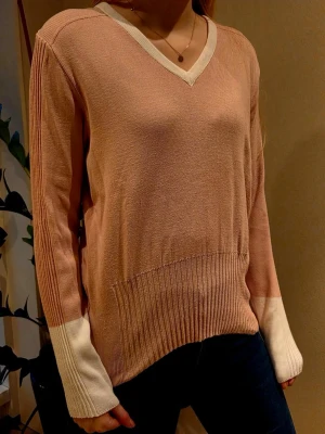 Stickad tröja i rosa från Ivanhoe - Snygg stickad tröja från Ivanhoe i ljusrosa med vita detaljer vid ärmslut och v-ringad hals. Tröjan har ribbstickad nederkant och muddar, samt en avslappnad passform. Perfekt för dig som gillar en stilren och enkel look.