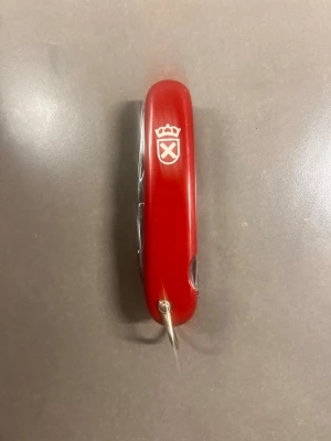 Röd multiverktygskniv med emblem - Säljer en röd multiverktygskniv med flera funktioner och ett vitt emblem med krona och korsade svärd på handtaget. Handtaget är i plast och verktygen i metall. Perfekt att ha med sig för olika småfix och äventyr.