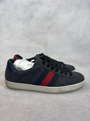 Gucci sneakers med blå och röda ränder - Snygga sneakers från Gucci i mörkblått med klassiska blå och röda sidoremsor. Skorna har diskret GG-mönster över hela ovandelen och svart snörning. Vit sula och detaljer i blått och rött på hälen. Perfekta för dig som gillar exklusiv streetstyle.