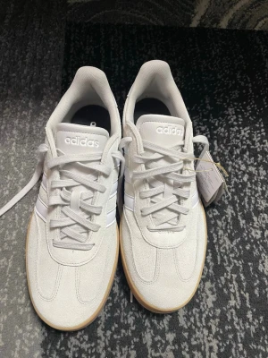 Vita Adidas sneakers med gummisula - Säljer ett par fräscha vita Adidas sneakers med klassiska tre ränder på sidan och ljusbrun gummisula. Skorna har lågt skaft, vita skosnören och ovandel i mockaliknande material. Perfekta för dig som gillar stilrena och sportiga skor.