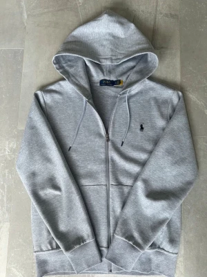 Grå zip hoodie Polo Ralph Lauren - Snygg grå hoodie med dragkedja från Polo Ralph Lauren. Perfekt för en avslappnad och stilren look. Tycker du om Polo Ralph Lauren så kommer du älska denna. 