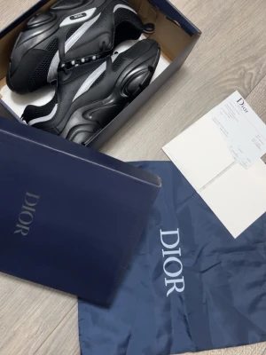 Svarta Dior sneakers med vita detaljer - Snygga svarta Dior sneakers med vita och grå detaljer. Skorna har chunky sula, meshpaneler och läderdetaljer för en modern look. Klassisk Dior-logga på sidan och bekväm innersula. Perfekta för dig som vill ha en exklusiv och trendig sneaker.
