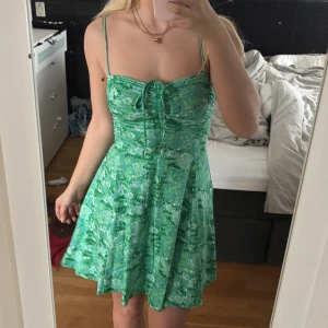 Grön blommig kortklänning med knyt - Snygg grön kortklänning med blommigt mönster och smala axelband. Klänningen har knyt framtill vid bysten och en lätt utsvängd kjol. Perfekt för varma dagar och somriga tillfällen.