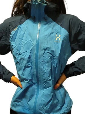 Blå vindjacka från Haglöfs - Snygg blå vindjacka från Haglöfs med mörkare blå partier på axlar och ärmar. Jackan har hög krage, hel dragkedja framtill och två sidofickor med dragkedja. Tillverkad i ett lätt och slitstarkt material, perfekt för blåsiga dagar.