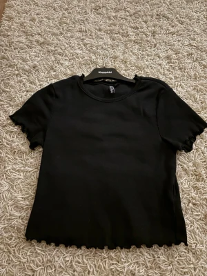 Svart ribbad topp från PIECES - Svart ribbad topp från PIECES med korta ärmar och vågiga kanter vid ärmslut och nederkant. Enkel och stilren design som passar till det mesta. Tillverkad i mjukt material för en bekväm känsla. Den känns som storlek S