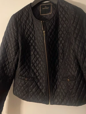 Svart quiltad jacka Soyaconcept - Stilren svart quiltad jacka från Soyaconcept med rund halsringning och dragkedja framtill. Jackan har två fickor med knappdetaljer och ett snyggt rutmönster över hela ytan. Perfekt för dig som gillar en klassisk men trendig look.