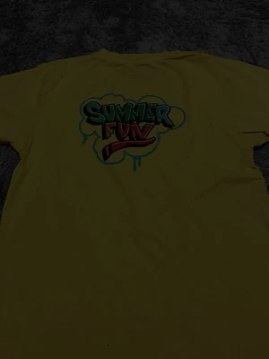 Gul t-shirt Summer Fun Stockholms stad - Gul t-shirt med färgglad 'Summer Fun'-grafik på ryggen och tryck av Stockholms stad på ärmen. Klassisk passform och korta ärmar, perfekt för sommardagar. Tillverkad i mjuk bomull för skön känsla.