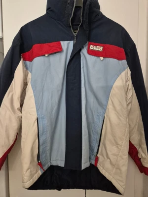 Skidjacka - Retro vindjacka från Gianiferoti i blått, ljusblått, vitt och rött. Jackan har hög krage, avtagbar huva och dragkedja med vindslå. Flera fickor framtill och cool färgblockad design. Tillverkad i slitstarkt syntetmaterial, perfekt för dig som gillar vintage och sportig look.