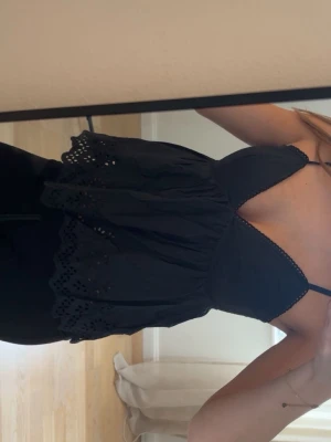 Svart broderad linnetopp med spets - Supersöt svart linnetopp med tunna axelband och broderade spetsdetaljer längs kanterna. Toppen har v-ringning och en luftig, lätt utställd passform med dekorativa hålbroderier längst ner.