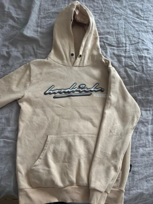 Beige hoodie från Hoodrich - Snygg beige hoodie från Hoodrich med stor logga i brun och vitt framtill. Har en liten svart fläck på luvan, se bild 5 och saknar snöre till luva