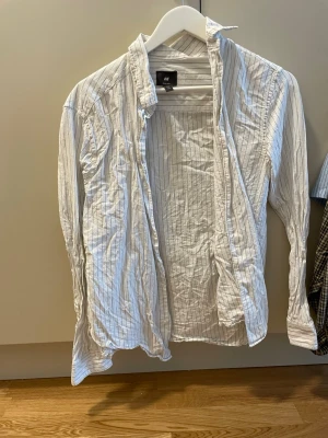 Vit randig skjorta från H&M - Vit skjorta från H&M med tunna grå ränder. Klassisk krage och långa ärmar. Skjortan är i lätt och luftig bomull, perfekt för en avslappnad stil. Passformen är normal och den har knappar framtill.