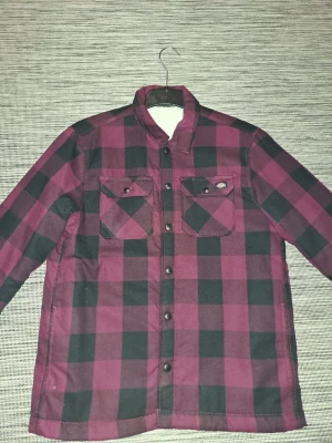 Rutig skjortjacka från Dickies - Snygg overshirt från Dickies i vinrött och svart rutigt mönster. Skjortjackan har tryckknappar framtill, två bröstfickor med lock och klassisk krage. Materialet är kraftigt och passar perfekt som lager på lager-plagg. Perfekt för dig som gillar streetwear och avslappnad stil.