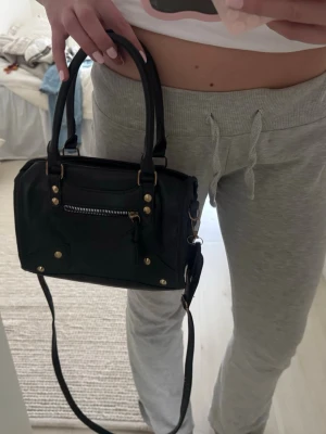 Svart handväska med gulddetaljer - Snygg svart handväska med både handtag och axelrem. Väskan har guldfärgade nitar och dragkedja framtill samt dekorativa sömmar. Perfekt storlek för det viktigaste och passar till många olika outfits.