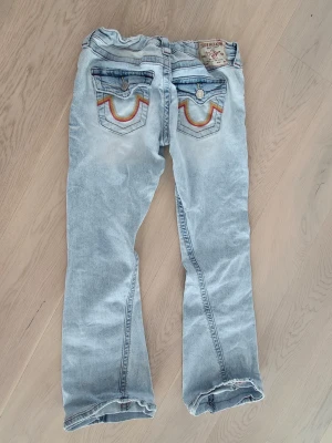 Ljusblå bootcut jeans True Religion, Joey low rise flare - Säljer ett par ljusblå bootcut jeans från True Religion med ikoniska broderade fickor bak i regnbågsfärger. Jeansen har låg midja. Originalstorlek 25. Uppsydda då dem var för långa och för stora vid midjan. Ej klippta och därför lätt att återställa.