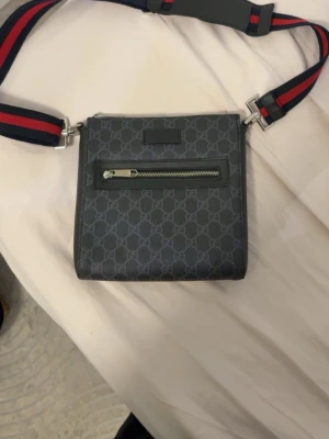 Gucci axelväska med GG-mönster - Snygg axelväska från Gucci i mörkgrått med klassiskt GG-mönster. Väskan har en justerbar axelrem i rött och marinblått, samt en praktisk ytterficka med dragkedja. Tillverkad i canvas med läderdetaljer och silverfärgade metalldelar.